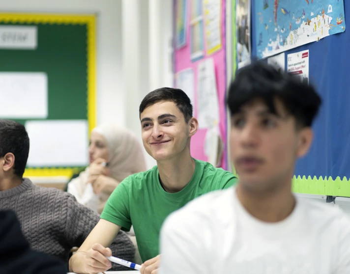 A young male student in a green t-shirt sits in a classroom, smiling while holding a whiteboard marker. Other students are visible in the background, including a student in a white hijab. A young male student in a green t-shirt sits in a classroom, smiling while holding a whiteboard marker. Other students are visible in the background, including a student in a white hijab.